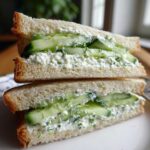 Two halves of a cucumber salad sandwiches stacked, showing creamy filling, fresh cucumber slices, and dill between white bread.