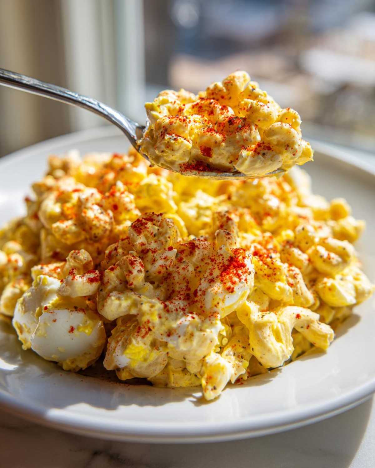 A spoonful of creamy deviled egg pasta salad, topped with paprika, being lifted from a white plate.