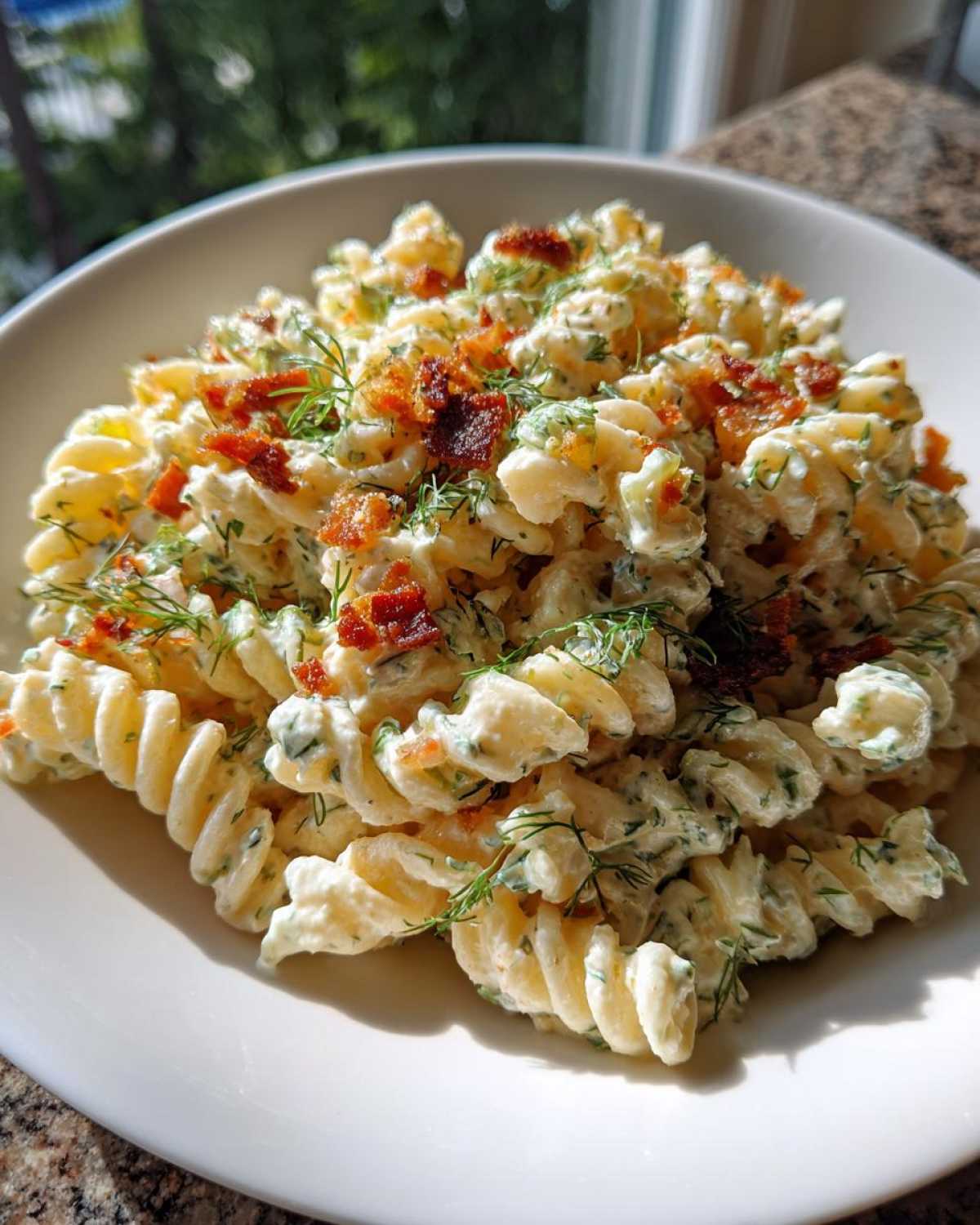 A close-up of creamy dill pickle bacon pasta salad made with rotini pasta, topped with crispy bacon bits and fresh dill.