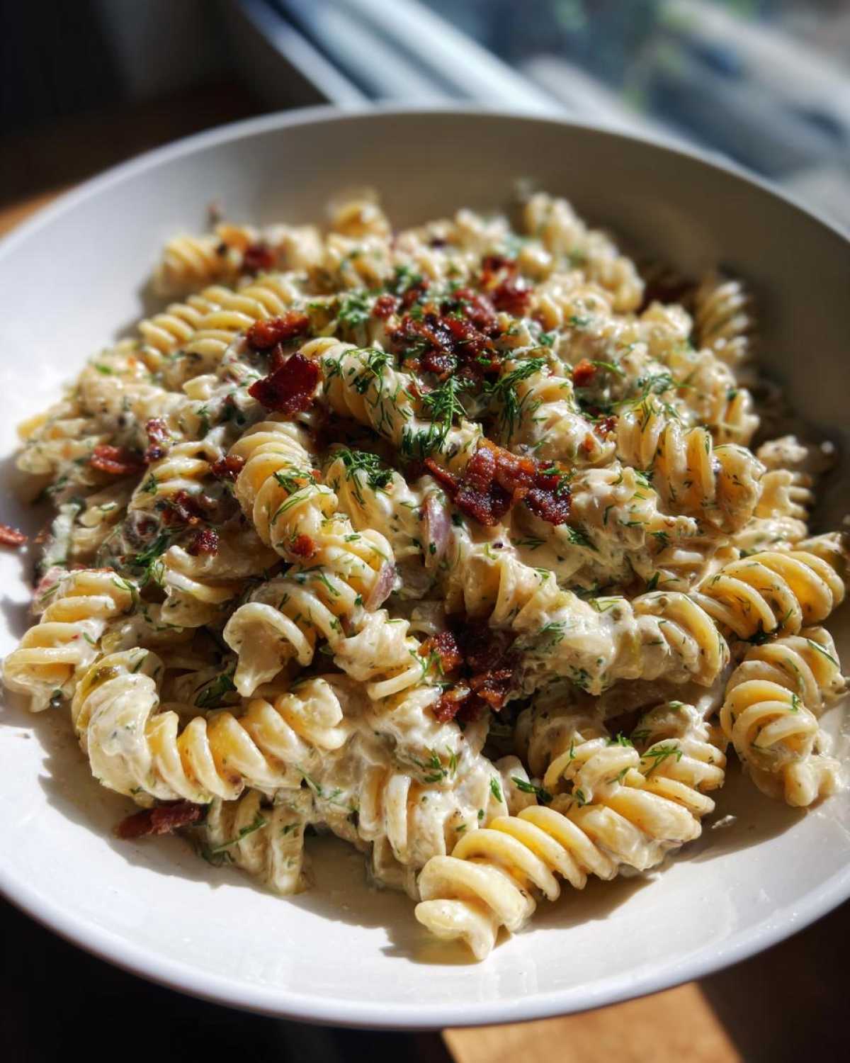 A close-up of creamy dill pickle bacon pasta salad made with rotini pasta, topped with crispy bacon and fresh dill.