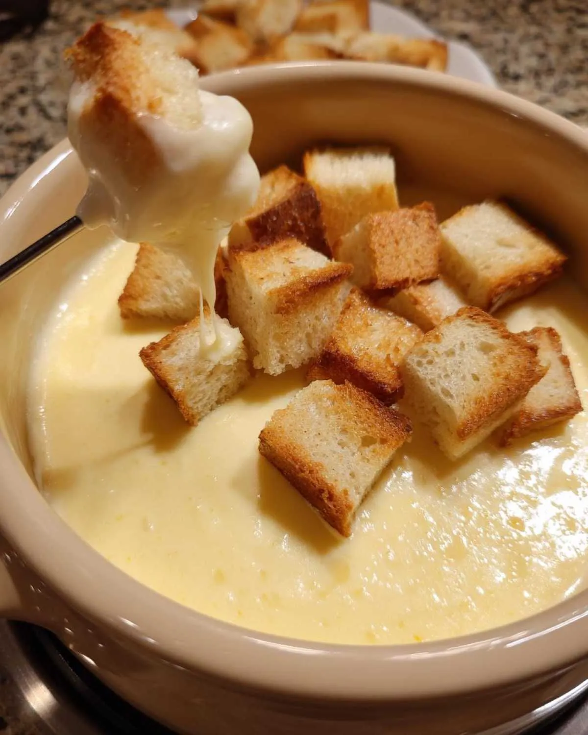 A piece of toasted bread is dipped into a warm pot of creamy cheese fondue recipe, dripping with melted cheese.