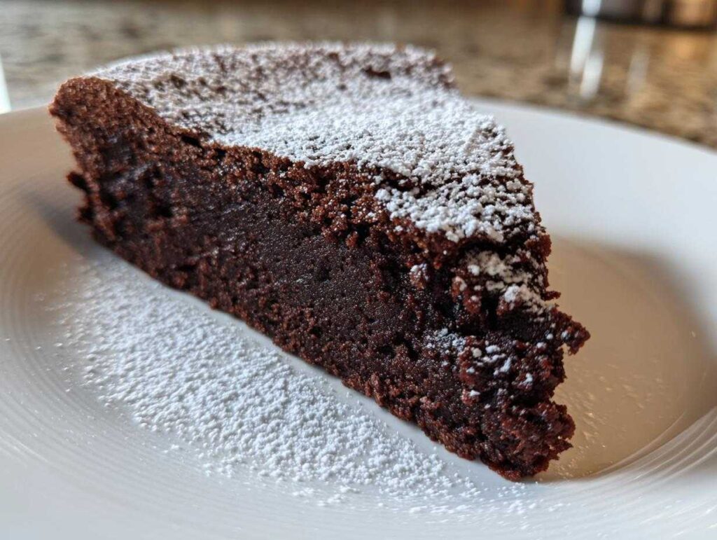 A close-up of a rich, dense slice of flourless chocolate cake dusted generously with powdered sugar on a white plate.
