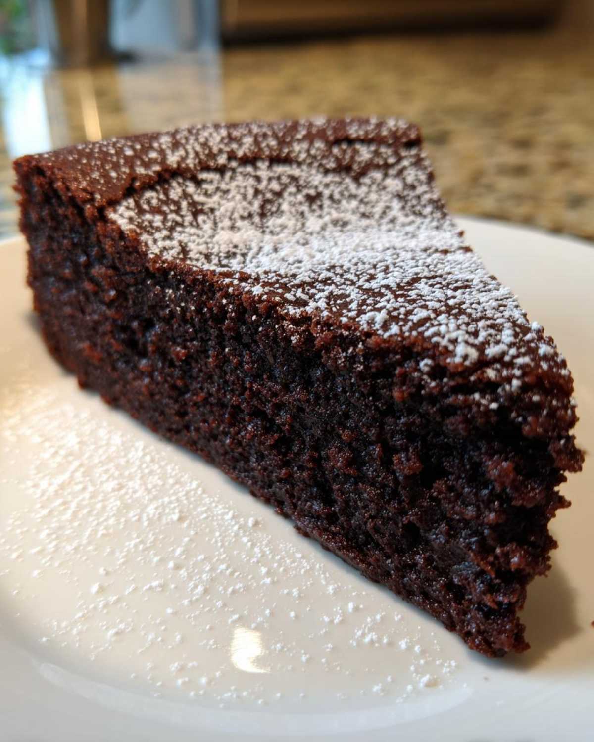 A close-up of a dark, moist slice of flourless chocolate cake dusted lightly with powdered sugar on a white plate.