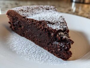 A close-up of a rich, dense slice of flourless chocolate cake dusted generously with powdered sugar on a white plate.