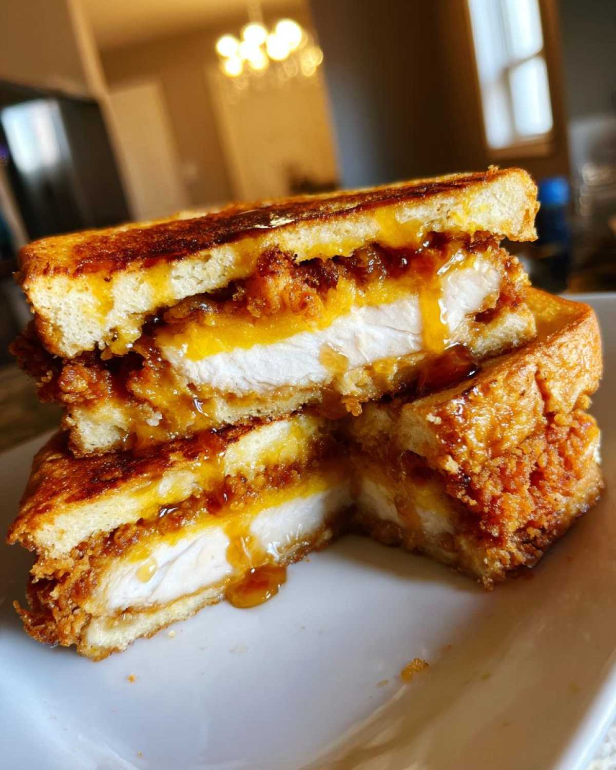 A close-up of a stacked french toast fried chicken sandwich cut in half, showing thick chicken breast, melted cheese, and dripping syrup.