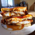 Close-up of a stacked french toast fried chicken sandwich, cut in half, drizzled generously with syrup.