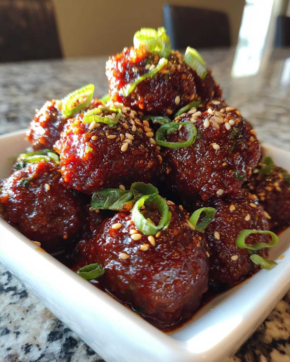 A close-up of glossy, glazed korean bbq meatballs piled high in a white bowl, topped with sesame seeds and sliced green onions.