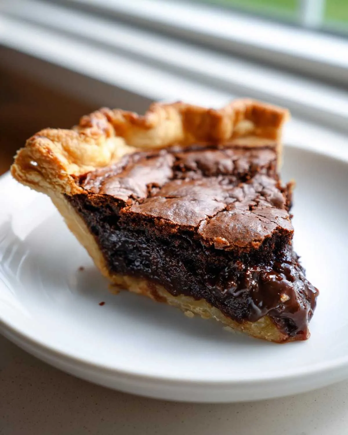 A close-up of a rich, gooey slice of fudge pie with a flaky crust, sitting on a white plate.