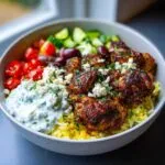 A close-up of a vibrant greek chicken rice bowl featuring seasoned chicken, yellow rice, tzatziki, tomatoes, cucumbers, and feta.