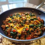 Close-up of a ground beef and spinach skillet topped with melted cheddar cheese and diced tomatoes.