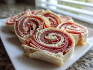 Close-up photo of several sliced Italian pinwheels featuring layers of salami, cheese, and tortilla on a white plate.