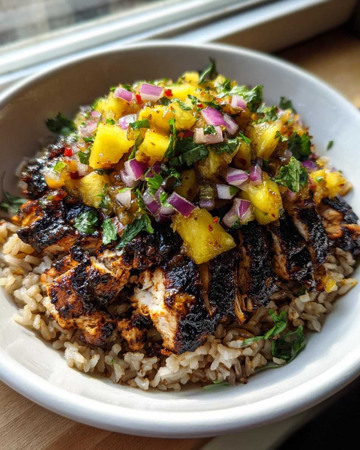 A close-up of a jerk chicken bowl featuring sliced, charred jerk chicken over brown rice, topped generously with bright pineapple salsa.