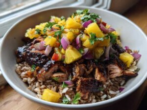 A close-up of a jerk chicken bowl featuring dark, glazed chicken pieces over brown rice, topped generously with bright pineapple salsa.