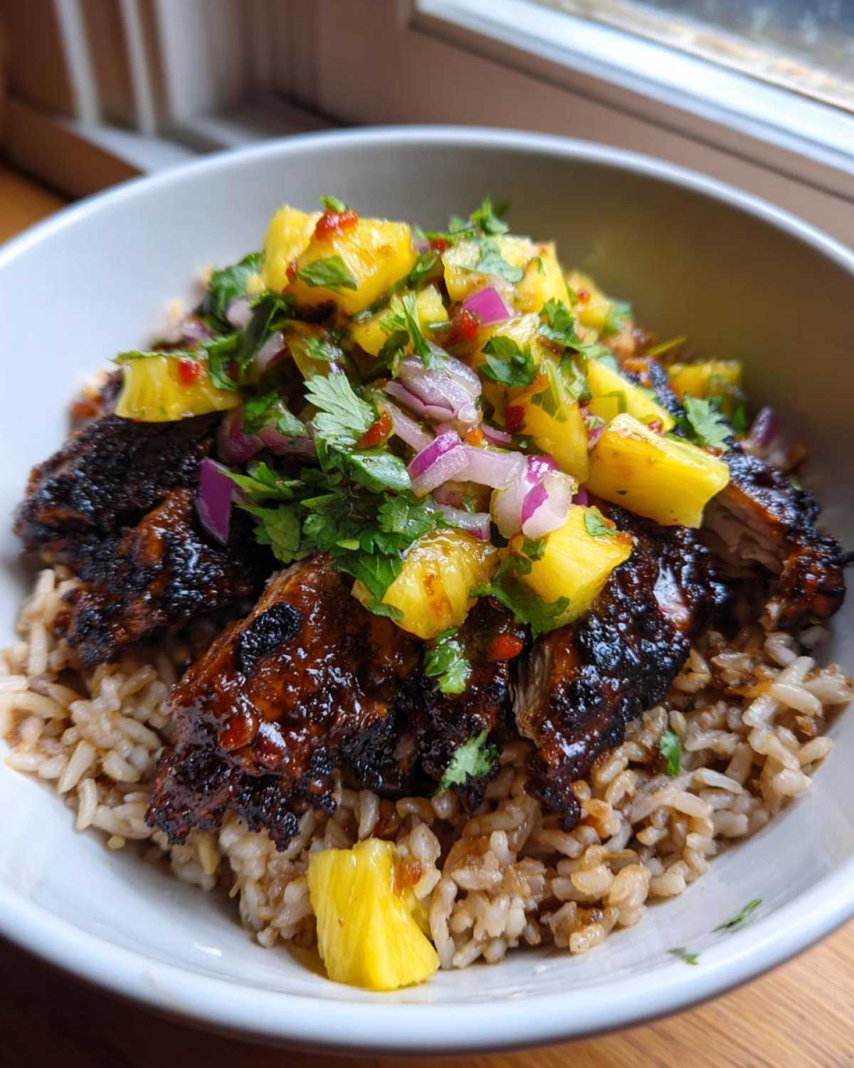 A close-up of a jerk chicken bowl featuring dark, glazed jerk chicken slices over rice, topped generously with bright pineapple salsa.
