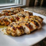 Close-up of sliced, juicy greek chicken breast seasoned heavily with herbs on a white plate.