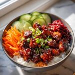 A close-up of a delicious korean bbq chicken bowls featuring glazed chicken pieces over rice, topped with sesame seeds, scallions, carrots, and cucumbers.
