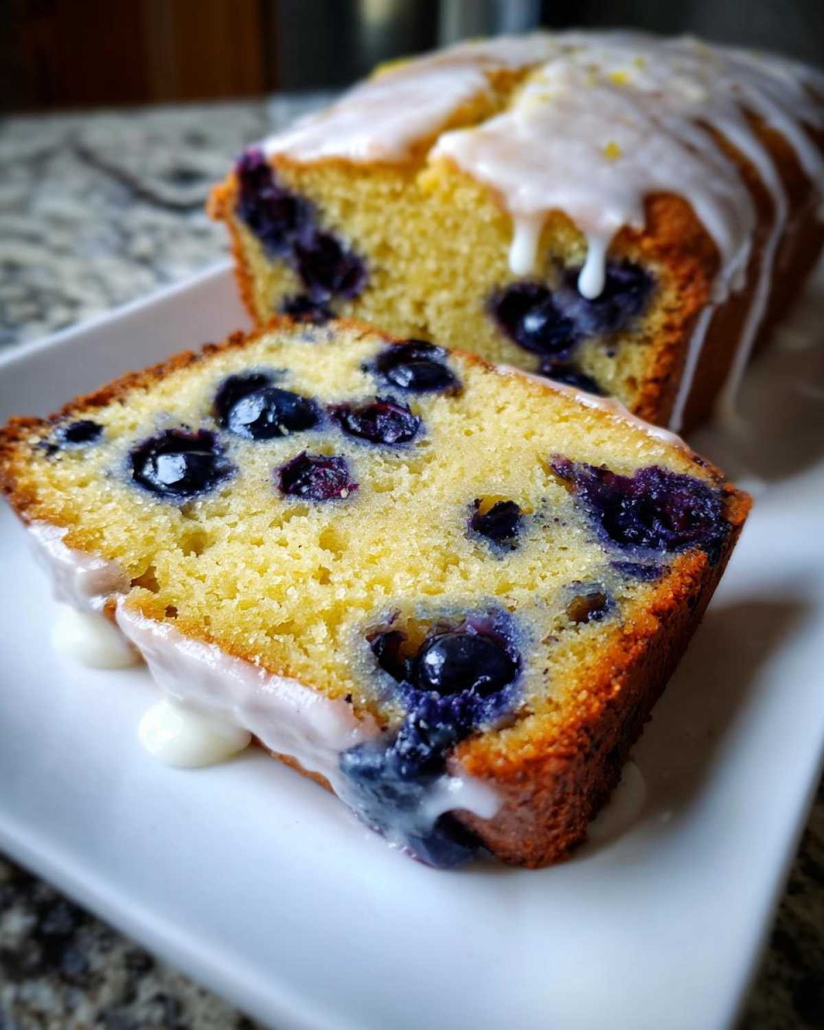 Close-up of a moist slice of lemon blueberry bread, topped with white glaze and full of blueberries.