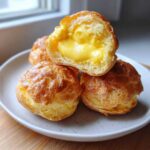 A stack of golden brown lemon cream puffs, one cut open to show the rich yellow cream filling.