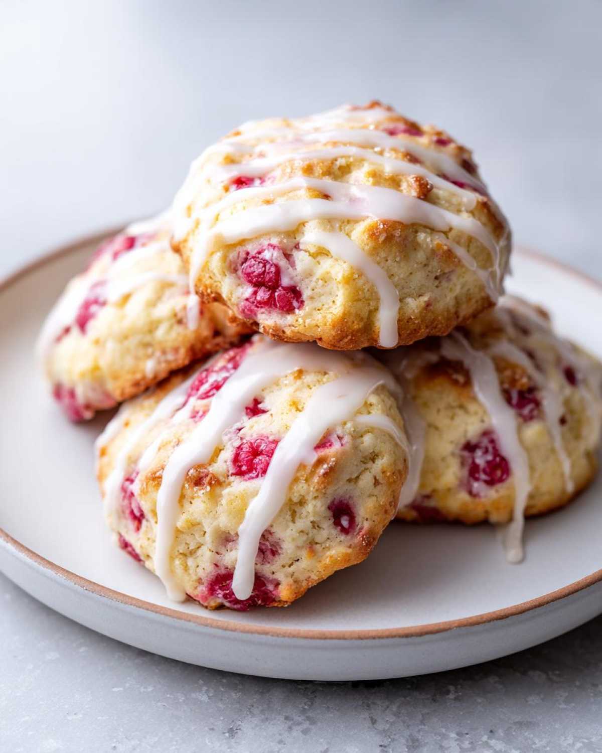 Close-up of four glazed lemon raspberry cookies stacked on a light plate, featuring visible raspberry pieces.
