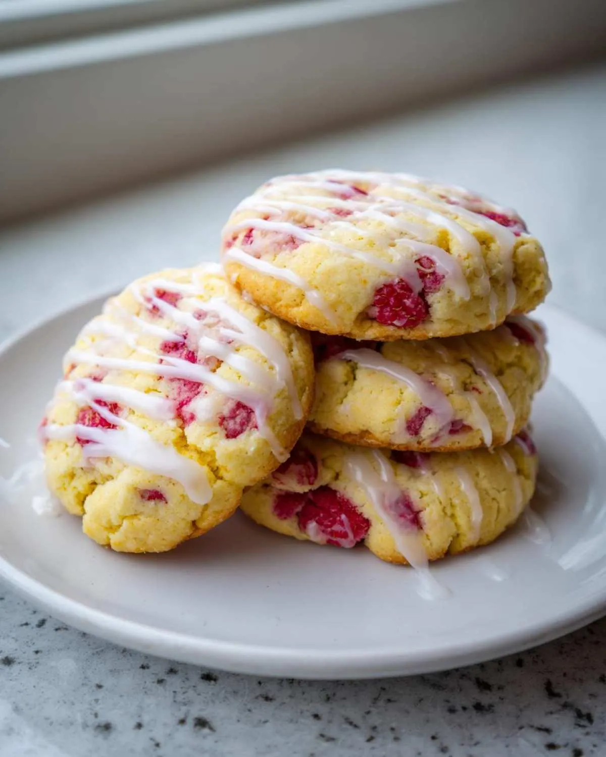 A stack of four soft lemon raspberry cookies drizzled with white lemon glaze, sitting on a white plate.