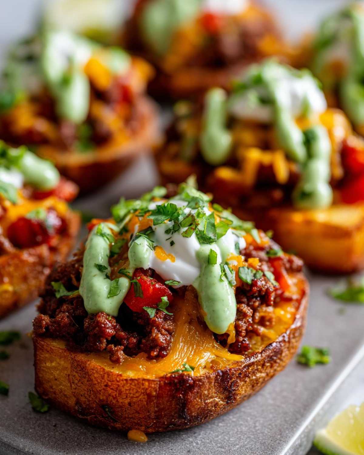 Close-up of a crispy potato half loaded with taco meat, melted cheese, sour cream, and green sauce, part of a loaded potato taco bowl.