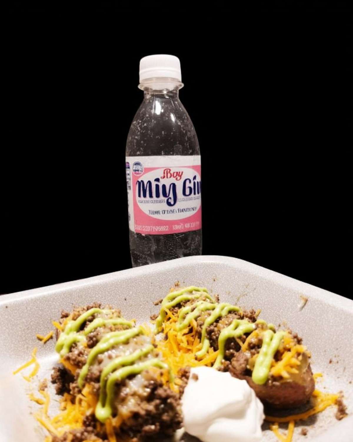 Close-up of a loaded potato taco bowl featuring seasoned ground beef, shredded cheese, green sauce drizzle, and sour cream, next to a bottle of Miy Gi soda.