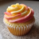 A single mango strawberry sunset cupcakes topped with swirled pink, yellow, and white sunset-colored frosting.