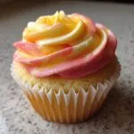A single mango strawberry sunset cupcakes topped with swirled pink, yellow, and white sunset-colored frosting.