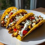 Three fully loaded mediterranean chicken tacos featuring seasoned chicken, feta, tomatoes, olives, and a white sauce on a white plate.