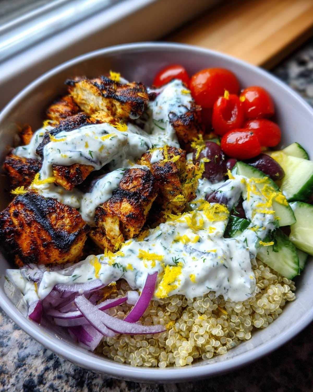 Close-up of a mediterranean lemon-dill chicken bowls featuring grilled chicken, quinoa, tzatziki sauce, cucumbers, tomatoes, and red onion.
