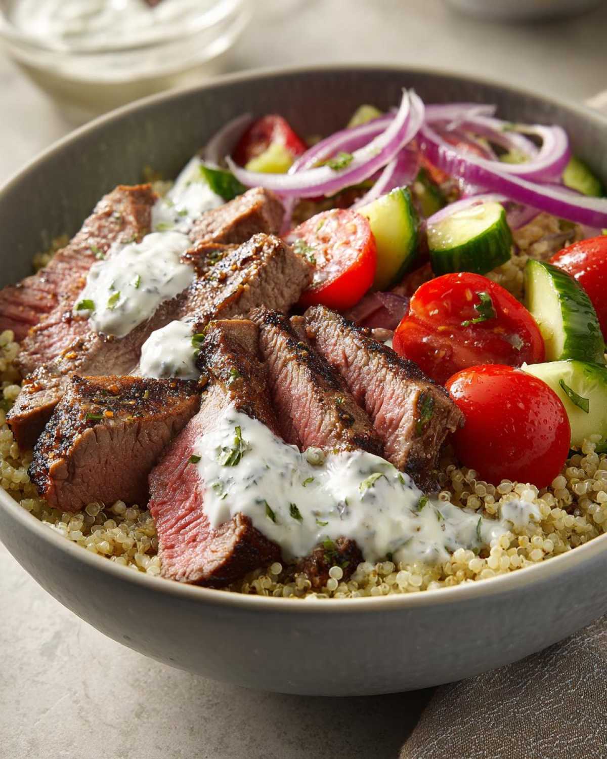 Close-up of a vibrant Mediterranean steak bowl featuring sliced medium-rare steak over quinoa, topped with cucumber, tomato, red onion, and tzatziki sauce.