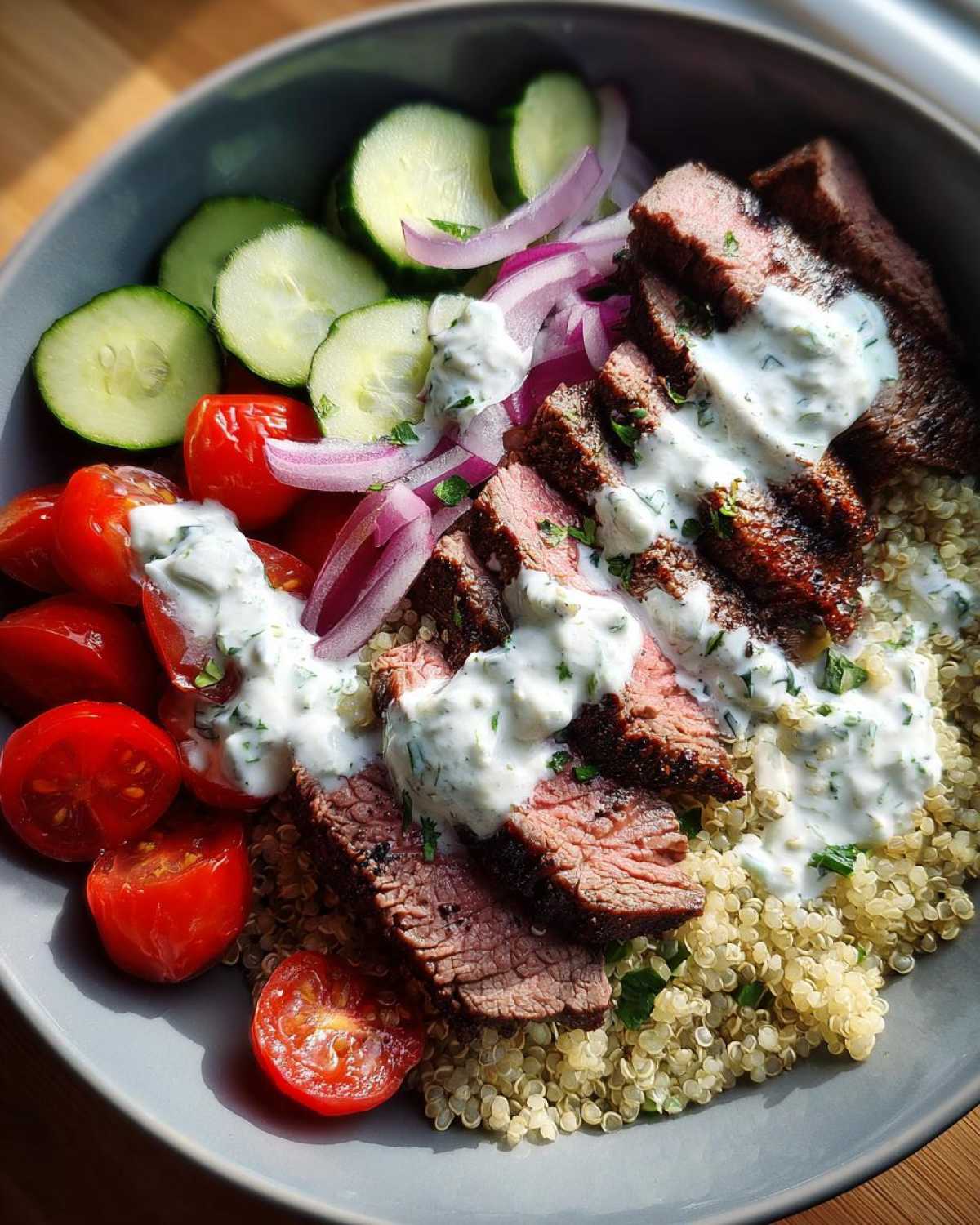 Close-up of a vibrant mediterranean steak bowl featuring sliced steak over quinoa, topped with tzatziki, cucumbers, tomatoes, and red onion.