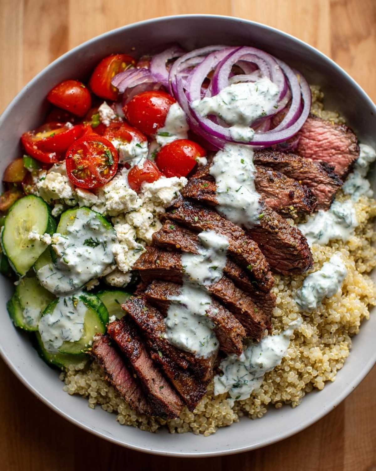Overhead view of a vibrant mediterranean steak bowl featuring sliced steak over quinoa, topped with tzatziki, feta, tomatoes, and red onion.