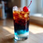 A tall glass of midnight malibu dirty soda showing distinct layers of white cream, bright blue syrup, and dark cola, topped with ice and a maraschino cherry.
