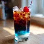 A tall glass of midnight malibu dirty soda showing distinct layers of white cream, bright blue syrup, and dark cola, topped with ice and a maraschino cherry.