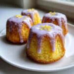 Four bright yellow mini lemon cakes with dripping lavender glaze and lemon zest topping.