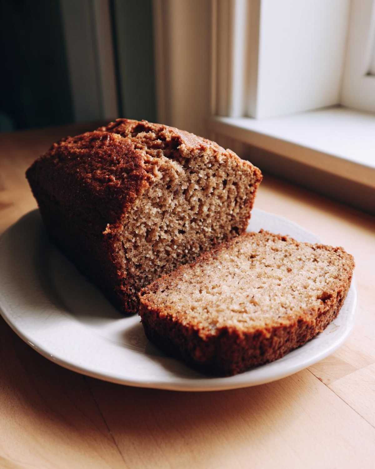A close-up of a moist banana bread recipe loaf, partially sliced, resting on a white plate near a window.