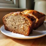 Two thick slices of moist banana bread recipe displayed on a white plate in bright sunlight.