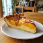 A close-up shot of a perfect slice of peach custard pie resting on a white plate on a wooden table.