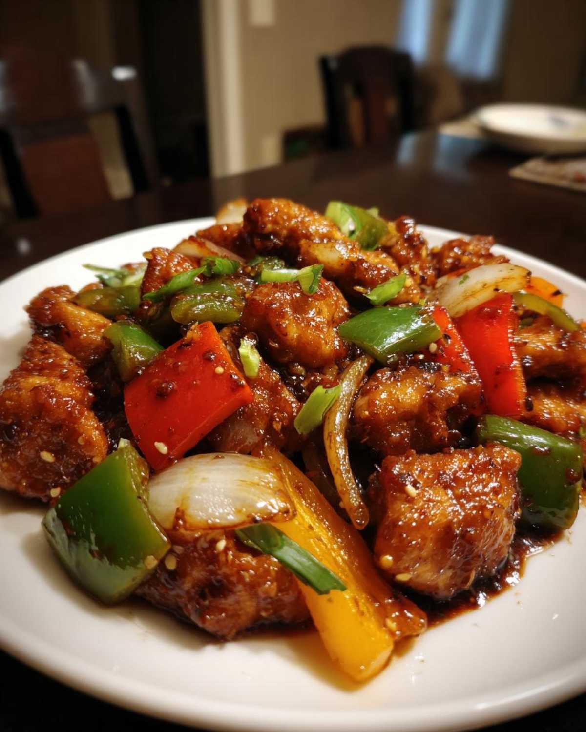A close-up shot of a serving of glossy pepper chicken stir fry featuring chunks of chicken coated in dark sauce with colorful bell peppers and onions.