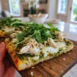 A close-up of a slice of pesto chicken flatbread topped with melted cheese, shredded chicken, and fresh arugula.