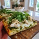 A close-up of a slice of pesto chicken flatbread topped with melted cheese, shredded chicken, and fresh arugula.