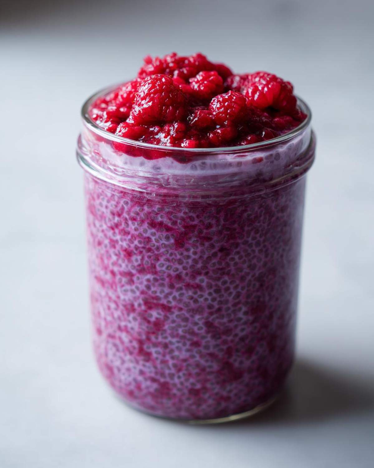 A glass jar filled with vibrant purple raspberry chia pudding, topped generously with fresh raspberries.