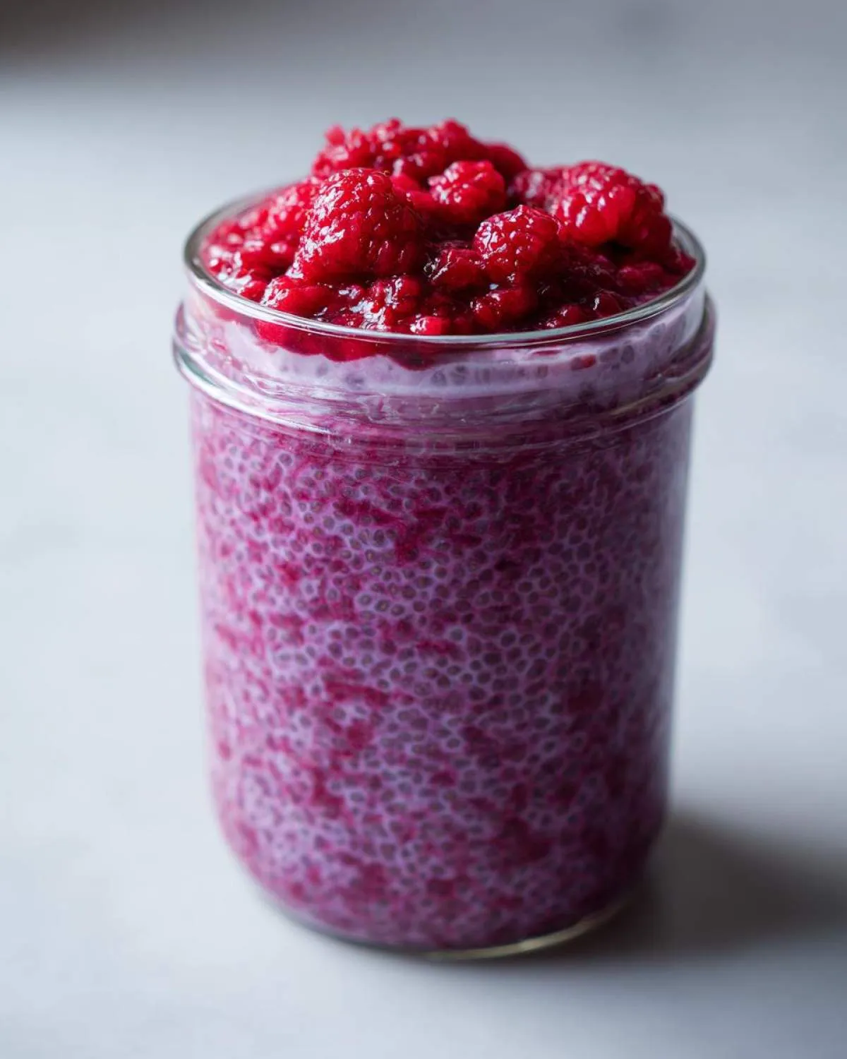 A glass jar filled with vibrant purple raspberry chia pudding, topped generously with fresh raspberries.