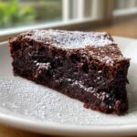 A decadent slice of flourless chocolate cake, dense and dark, dusted generously with powdered sugar on a white plate.