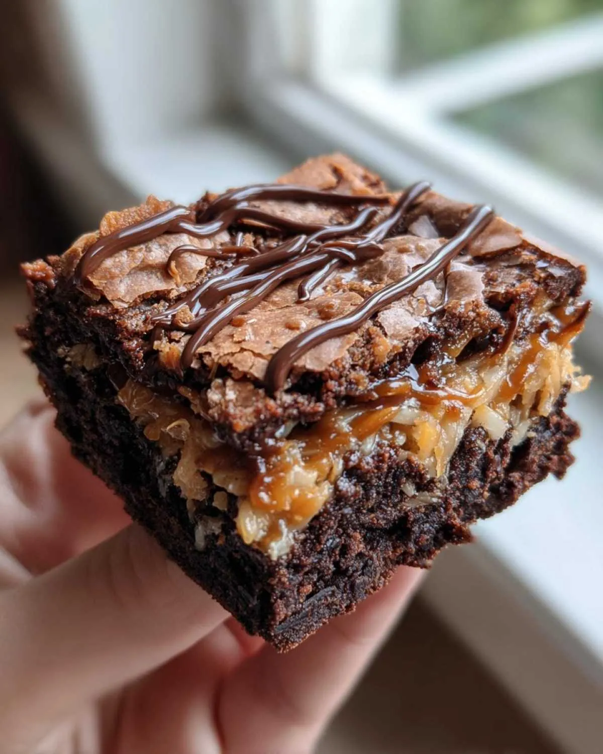 A hand holding a rich, fudgy square of samoa brownies topped with caramel, coconut, and a chocolate drizzle.