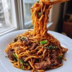 A close-up of saucy korean beef noodles being lifted high from a white bowl with chopsticks, garnished with sesame seeds.