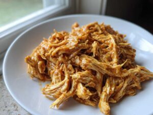 Close-up of shredded slow cooker honey mustard chicken coated in a savory sauce, served on a white plate.