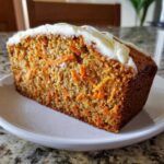A close-up of a thick slice of carrot cake banana bread, featuring visible shredded carrots and topped with white cream cheese frosting.