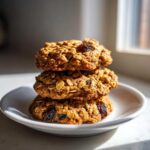 A stack of three chewy oatmeal raisin cookies resting on a small white plate, highlighted by natural window light.
